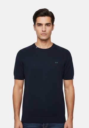 Uomo che indossa una maglietta a maniche corte in maglia di colore blu navy scuro con un piccolo logo geometrico turchese sul petto, in piedi davanti a uno sfondo neutro.