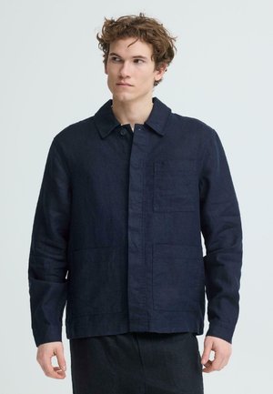 Junger Mann mit lockigem Haar, der eine dunkelmarinefarbene Jacke mit drei Vordertaschen trägt, vor einem schlichten hellen Hintergrund stehend.