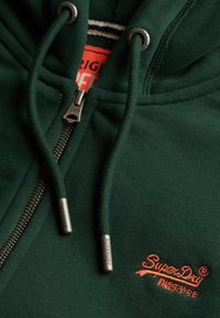 Donkergroene hoodie met ritssluiting en trekkoorden met metalen uiteinden, een gedeeltelijk zichtbaar oranje label aan de binnenkant, en een oranje SuperDry-logo geborduurd.