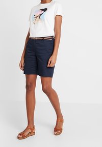 Bílé tričko s grafickým designem, tmavě modré šortky a hnědé sandály. Oblečení zahrnuje hnědý opasek a má uvolněný střih.