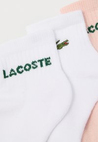 Τρία ζευγάρια καλτσών: ένα λευκό με πράσινο λογότυπο "LACOSTE" και κροκόδειλο, ένα ροζ και ένα λευκό. Μείγμα από βαμβάκι με ραμμένα λάστιχα.
