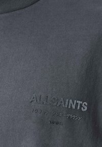 Dunkelgrauer Stoff mit erhabener Stickerei des Textes "ALLSAINTS" und darunter kleinerem Text in einer anderen Sprache, zentriert auf der Textiloberfläche.