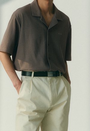 Man draagt een bruin overhemd met korte mouwen, gestopt in beige geplooide broek met een zwarte riem, handen in de zakken.