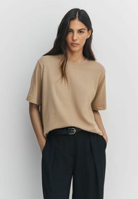 Beżowa koszulka z krótkim rękawem wykonana z miękkiego materiału. Luźny krój, okrągły dekolt. Dopasowana do wysokiej talii czarnych spodni i ciemnego paska.