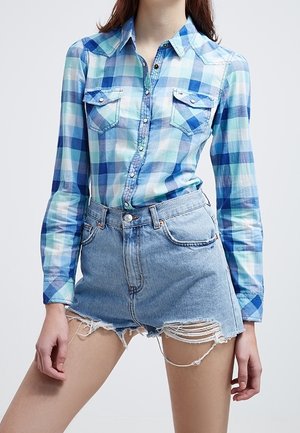 Personne portant une chemise à carreaux bleue et blanche boutonnée, rentrée dans un short en denim clair usé.