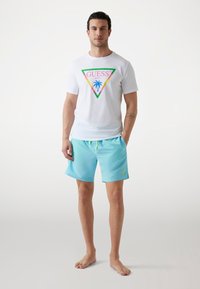 Hvid T-shirt med multicoloreret trekantet grafik, der har "GUESS BEACH" og et palmetræ. Lyseblå shorts med snøre og logo.