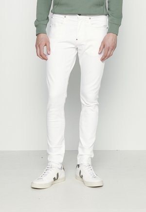 Mand iført hvidt slim-fit jeans, hvide sneakers med sort logo og en grøn langærmet top, stående foran en ensartet lys baggrund.