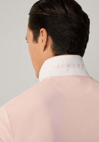 Polo tričko s texturovaným tělem v světle růžové barvě a bílým límečkem. Na límečku je jemný růžový nápis značky "HACKETT."