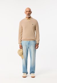 Beige tröja med v-ringning, grön logotyp, ljusgul skjorta med krage, ljusblå denimjeans, håller en beige keps, krämfärgade sneakers med bruna sulor.