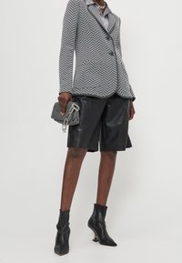 Blazer gris con patrón de espiga y cierre de dos botones, combinado con pantalones cortos de cuero negro brillantes y botas de tobillo negras con tacón, sosteniendo un bolso gris.