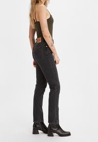 Mujer con cabello largo y rubio, vestida con camiseta sin mangas negra a rayas, jeans negros de talle alto y botines negros, de pie en pose lateral sobre fondo blanco.