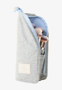 Grå teksturert sminkepenselveske med en lys blå glidelås. Inneholder flere pensler med myke bust, med en etikett foran.