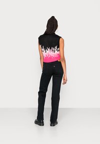 Top de malha sem mangas preto com padrão de chamas em rosa e branco, combinado com jeans pretos de perna reta e sapatos pretos, visto de costas.