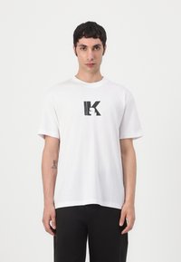 T-shirt branca de algodão com mangas curtas, apresentando um logótipo estilizado em preto da letra "K" impresso ao centro. Corte simples e descontraído, com textura suave.