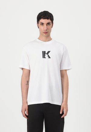 White cotton t-shirt with short sleeves, featuring a black stylized "K" logo printed centrally. Simple, relaxed fit, soft texture.