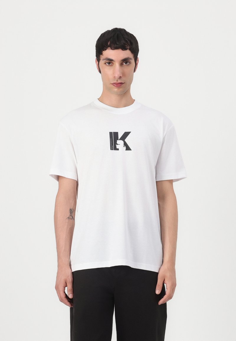 T-shirt branca de algodão com mangas curtas, apresentando um logótipo estilizado em preto da letra "K" impresso ao centro. Corte simples e descontraído, com textura suave.
