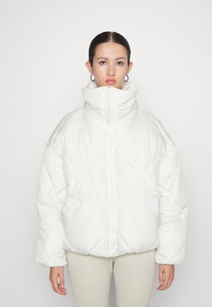 Jeune personne portant une veste matelassée blanche à col montant zippée, associée à un pantalon beige clair à côtes.