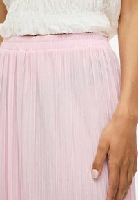 Gonna plissée rosa con vita elasticizzata, realizzata in tessuto leggero, con una consistenza morbida e una leggera lucentezza. Mano appoggiata sulla gonna.