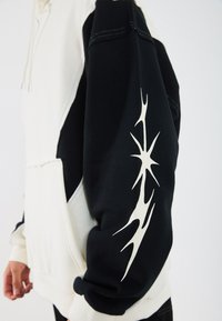 Zwart-wit hoodie met lange mouwen. De zwarte mouw heeft een abstract ivoorkleurig ontwerp. Zachte stof met een kangoeroezak aan de voorkant.