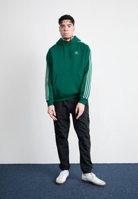 Sweatshirt verde com riscas laterais, bolso canguru frontal e logótipo bordado. Usado com calças cargo pretas e ténis brancos.