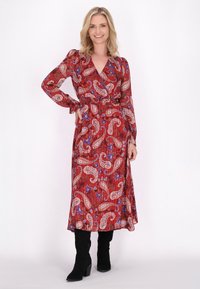 Rotes Kleid mit Paisleymuster, langen Ärmeln, Wickelausschnitt und taillierter Silhouette, kombiniert mit kniehohen schwarzen Wildlederstiefeln.