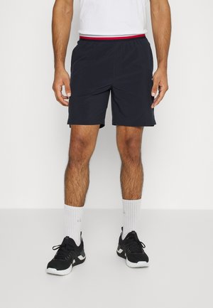 Mann trägt schwarze Sportshorts mit einem rot-weiß-blauen Bund, weiße Socken und schwarze Laufschuhe, steht vor weißem Hintergrund.