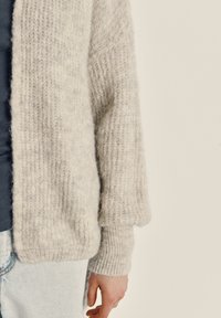 Svetlo sivi pleteni pulover z ohlapnimi rokavi, rebrastimi manšetami in mehkim, teksturiranim zaključkom. Nošen preko temne srajce s modrimi hlačami iz jeansa.