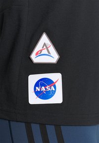 Černé tričko s dvěma nášivkami: trojúhelníková nášivka s červenými a modrými motivy a čtvercové logo NASA na bílém pozadí.