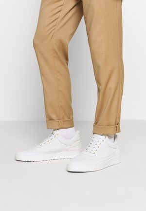 Person iført beige bukser med manchetter og hvide snøresneakers med hvide sokker på en ensfarvet hvid baggrund.