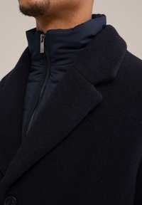 Marineblauwe wollenmix overjas met hoge kraag en een ritssluiting aan de binnenkant. Heeft een soepele textuur en knoopsluiting.