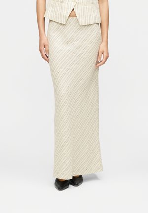 Jupe beige à rayures ondulées verticales de couleur marron clair, longue et ajustée, accompagnée d'un gilet assorti. Des chaussures noires complètent la tenue.