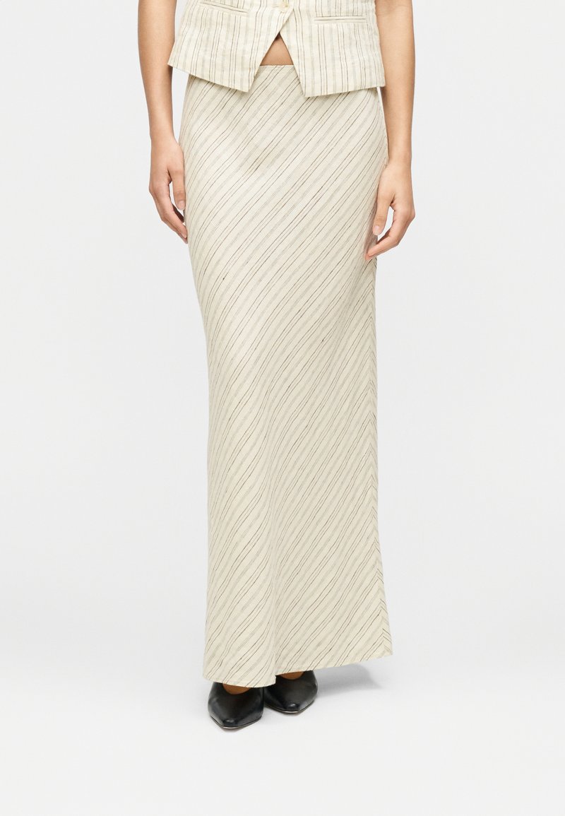 Μπεζ φούστα με κάθετες κυματιστές ρίγες σε ανοιχτό καφέ, μακριά και εφαρμοστή, συνδυασμένη με γιλέκο σε αντίστοιχο χρώμα. Μαύρα παπούτσια ολοκληρώνουν την εμφάνιση.