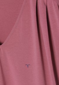 Růžový top bez rukávů s kulatým výstřihem, vyrobený z měkké tkaniny; má malé logo v tmavě fialové barvě na přední straně a drapérii.