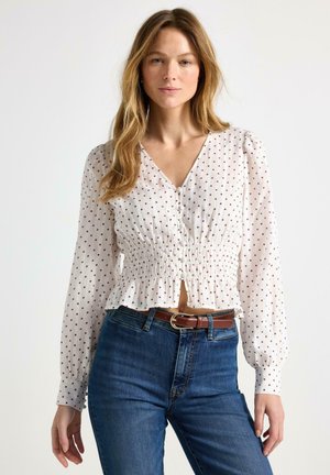Donna che indossa una camicetta bianca a maniche lunghe con pois rossi, vita arricciata, bottoni frontali, abbinata a jeans blu e cintura marrone.