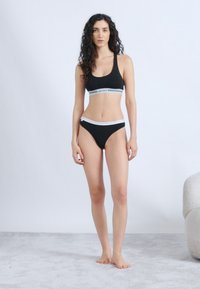 Woman with curly hair stands barefoot wearing black sports bra and underwear with white elastic bands against plain light background.