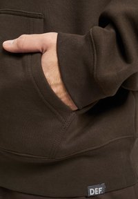 Brun genser med kengurulomme, ribbede ermer og nederkant. En liten etikett med "DEF." er synlig nær bunnen.