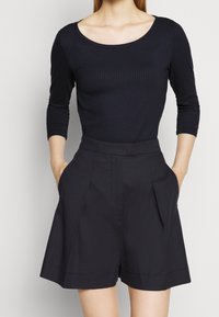 Frau in einem schwarzen gerippten Shirt mit drei Viertel Ärmeln und hochgeschnittenen schwarzen Shorts, mit den Händen in den Taschen, von Hals bis zur Mitte des Oberschenkels dargestellt.