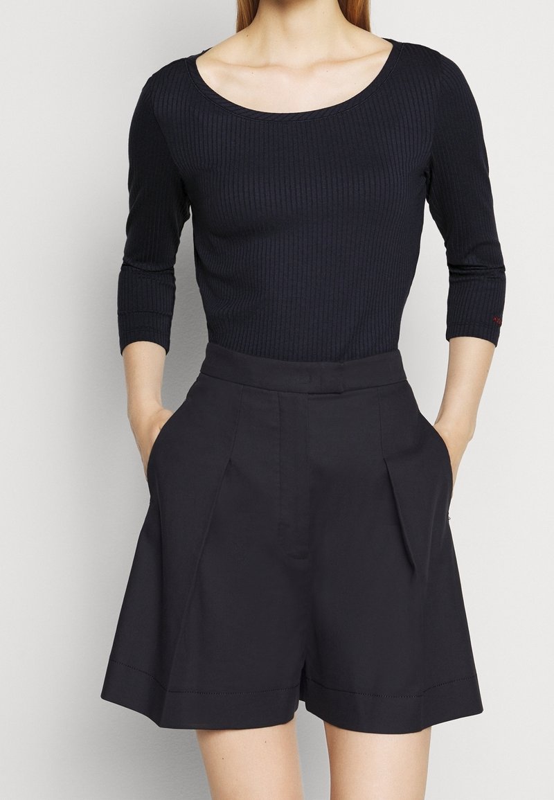 Frau in einem schwarzen gerippten Shirt mit drei Viertel Ärmeln und hochgeschnittenen schwarzen Shorts, mit den Händen in den Taschen, von Hals bis zur Mitte des Oberschenkels dargestellt.