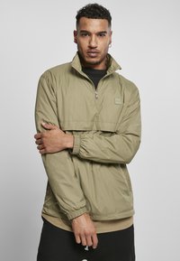 Letvægts olivengrøn pulloverjakke med halv-zip krave, elastiske manchetter og en forlomme, båret over en beige skjorte og sorte bukser.