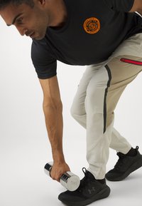 T-shirt noir avec logo orange, pantalon de sport beige avec fermeture éclair rouge et accents gris, tenant un haltère en argent, portant des baskets noires.