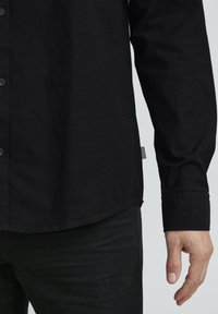 Black button-up shirt with a smooth texture, long sleeves, and a curved hem. A small label with a striped pattern is visible on the side.