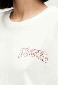 Κοντινό πλάνο ενός ατόμου που φοράει λευκό πουκάμισο με κεντητό κόκκινο λογότυπο "DIESEL" στο στήθος.