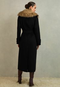 Garš melns mētelis ar mākslīgās kažokādas apkakli, jostu viduklī un strukturētām piedurknēm. Svārki sasniedz zem ceļgaliem; valkāti ar brūnām papēžu zābakiem.