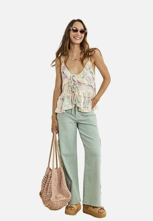 Mujer con blusa floral con volantes, vaqueros de pierna ancha color verde claro, sandalias de plataforma, gafas de sol, collar y bolso grande de mimbre.