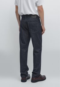 Dark blue denim jeans with a relaxed fit, featuring a straight leg, two back pockets, and a plain black waistband tag. Worn with brown shoes.