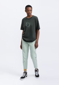 Donkergroene katoenen t-shirt met een grafisch ontwerp, gecombineerd met licht muntgroene taps toelopende broek. Zwarte hoge sneakers met witte accenten.