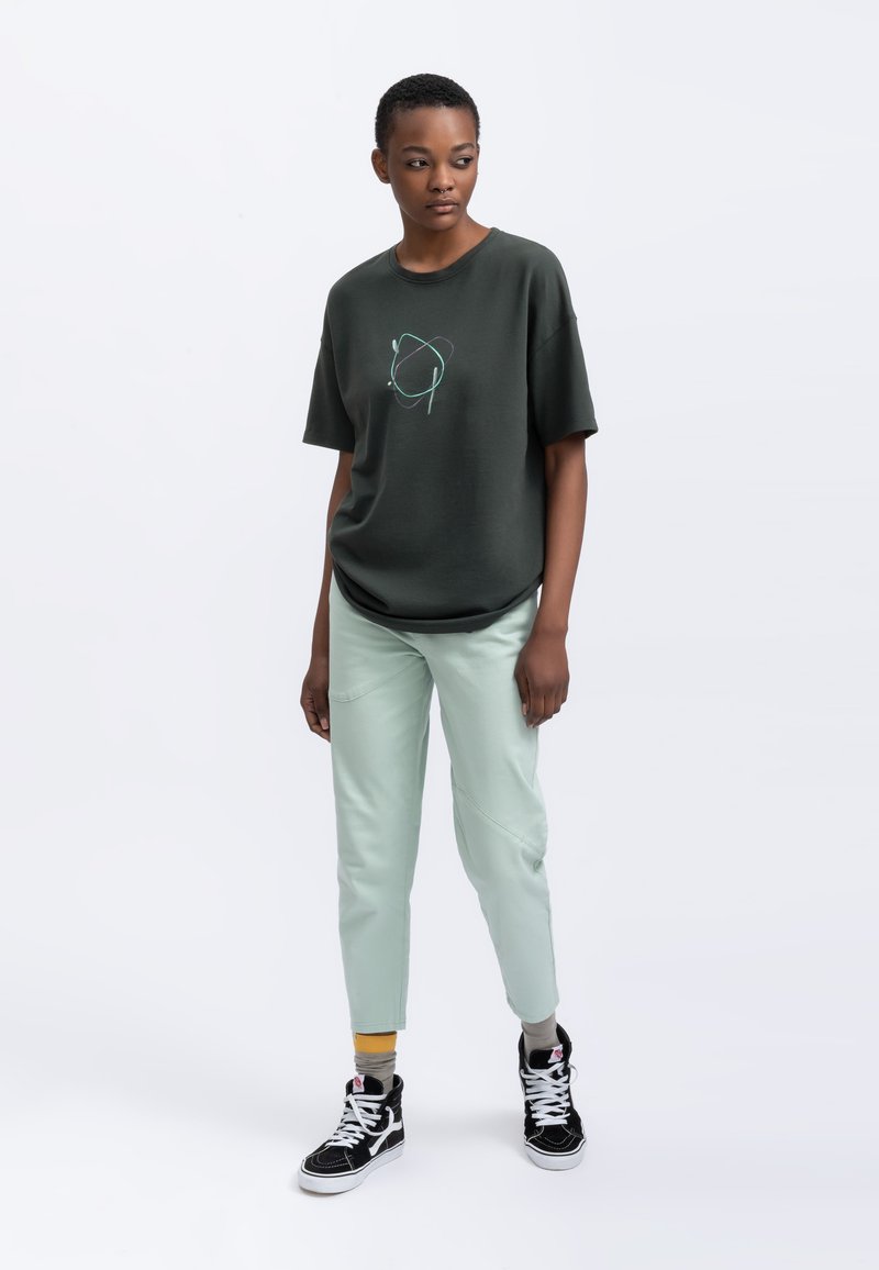 Donkergroene katoenen t-shirt met een grafisch ontwerp, gecombineerd met licht muntgroene taps toelopende broek. Zwarte hoge sneakers met witte accenten.