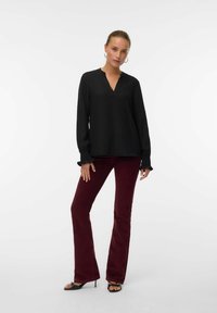 Donna che indossa una blusa nera a maniche lunghe con polsini a volant, pantaloni bordeaux, sandali neri con tacchi alti e orecchini a cerchio, in piedi su uno sfondo bianco.