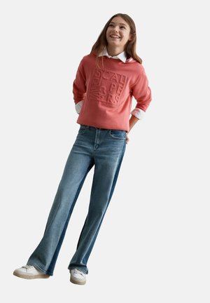 Joven sonriente, lleva un suéter coral sobre una camisa blanca, vaqueros azules de pernera ancha y zapatillas blancas, de pie con las manos detrás de la espalda.