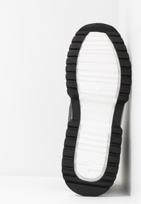 Černá gumová podrážka s bílými akcenty, texturovanými hřebeny a hladkou střední oblastí. Na spodní straně je logo značky.